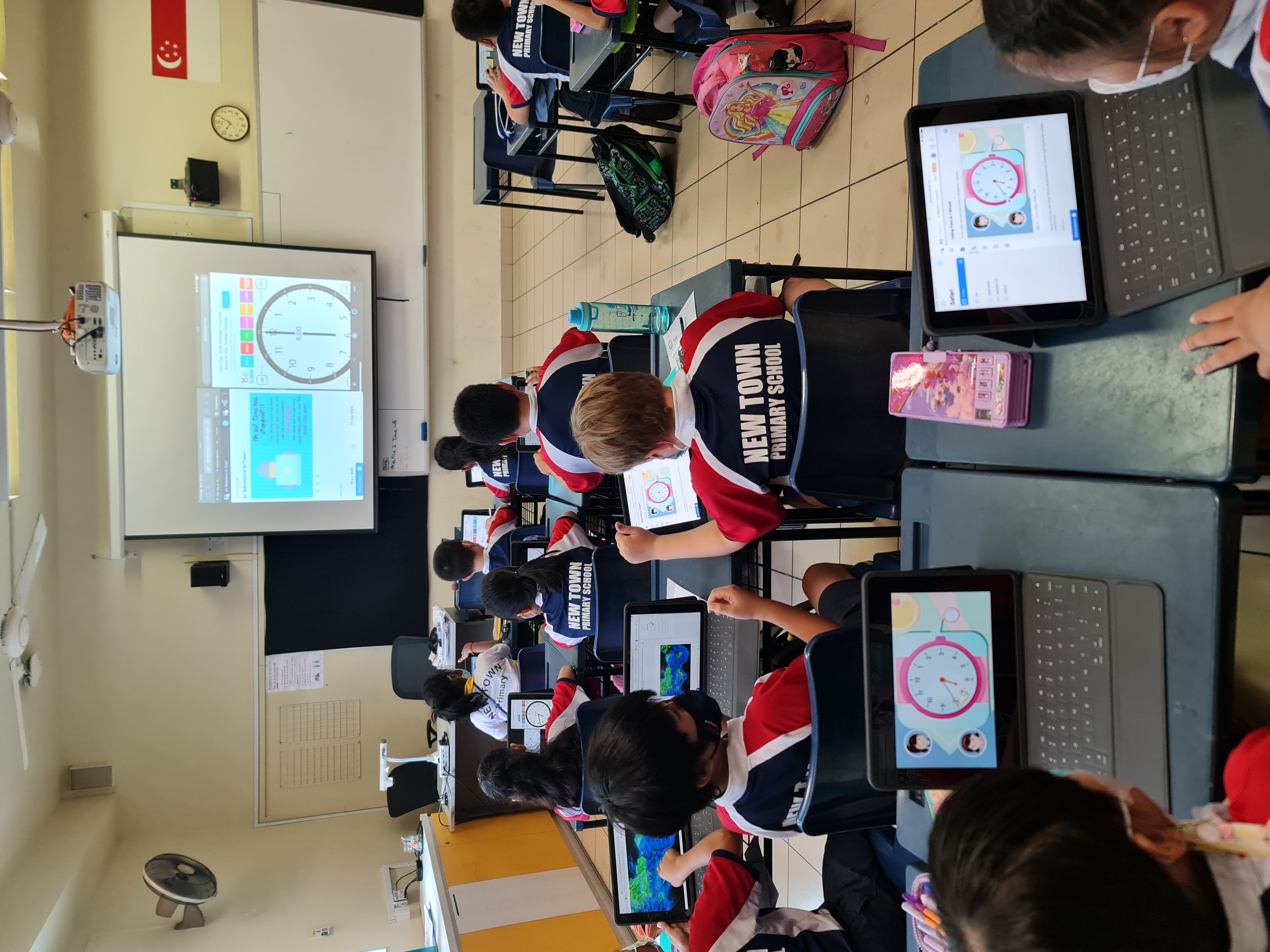 New Town Primary School students sit at desks with tablets showing a clock lesson on a projected screen.